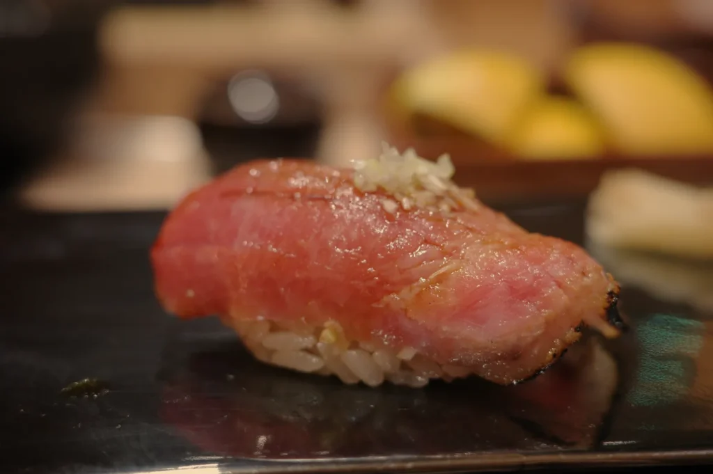 A piece of nigiri sushi topped with a slice of seared tuna, garnished with finely chopped garlic, served on a dark plate.