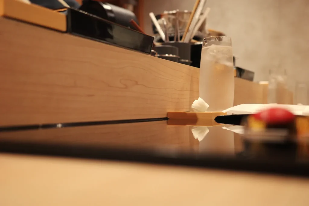 A wooden bar with a clear glass of water and a small piece of wasabi on a plate, with soft focus on the foreground.