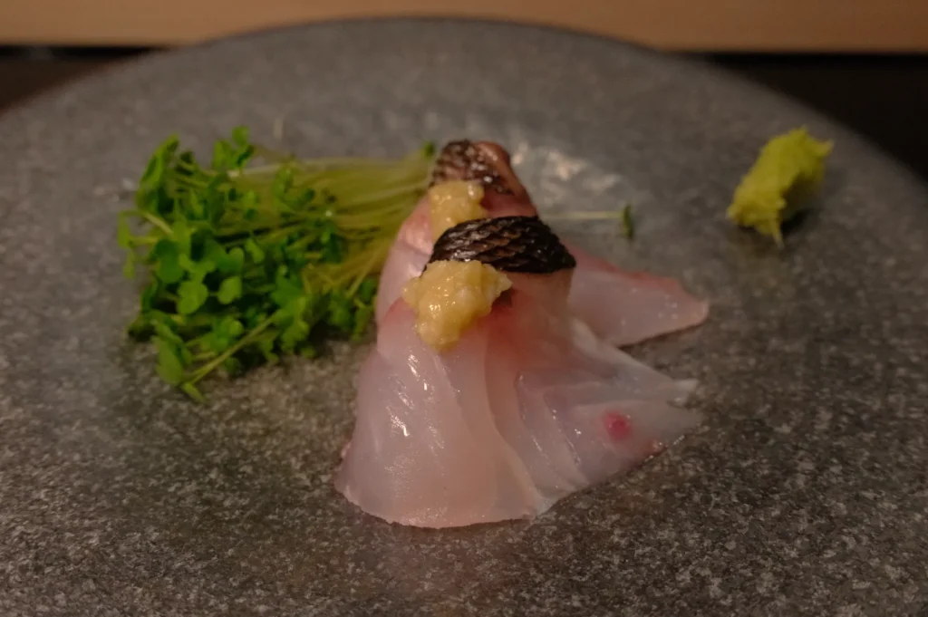 A plate with two pieces of sliced fish topped with a small amount of sauce, accompanied by greens and a piece of wasabi.