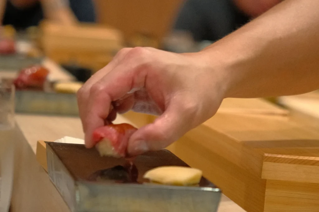 A hand picking up sushi from a metal serving tray in a restaurant setting.