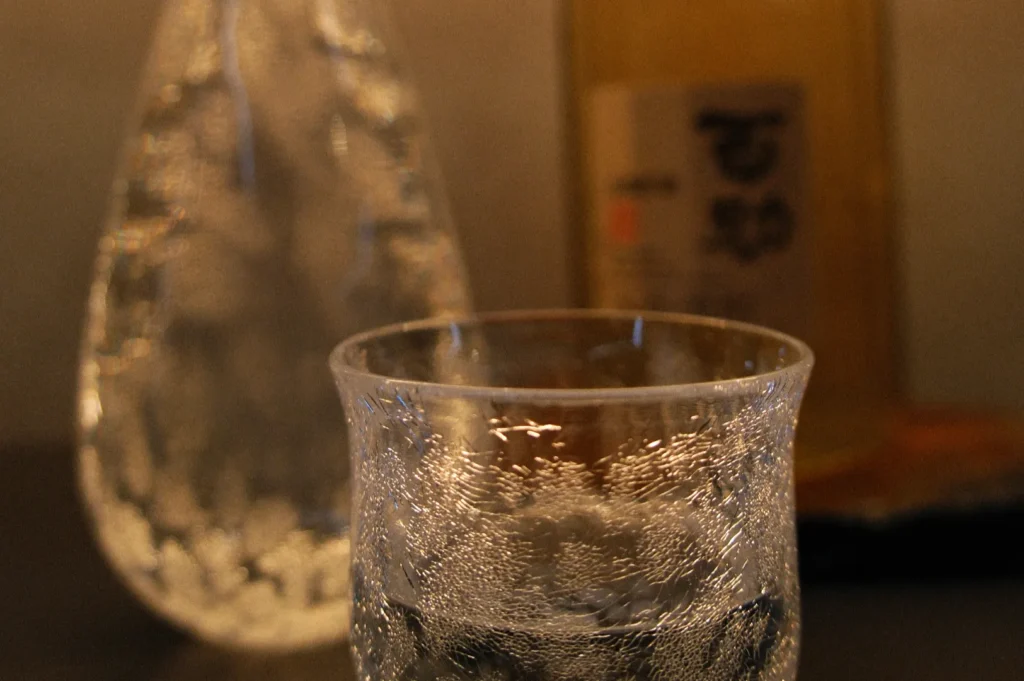 A glass of water with a textured surface in the foreground, with a decanter and a bottle in the background.
