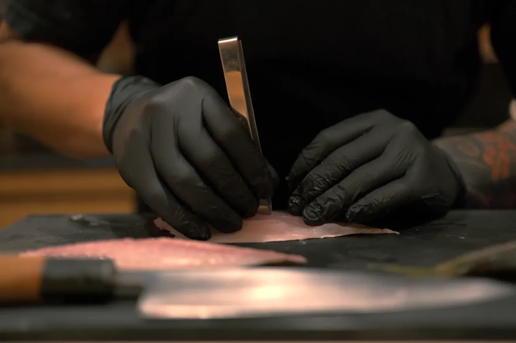 Chef Masa with pair of gloved hands using a tool to prepare fish fillets on a cutting board, with knives visible in the foreground.