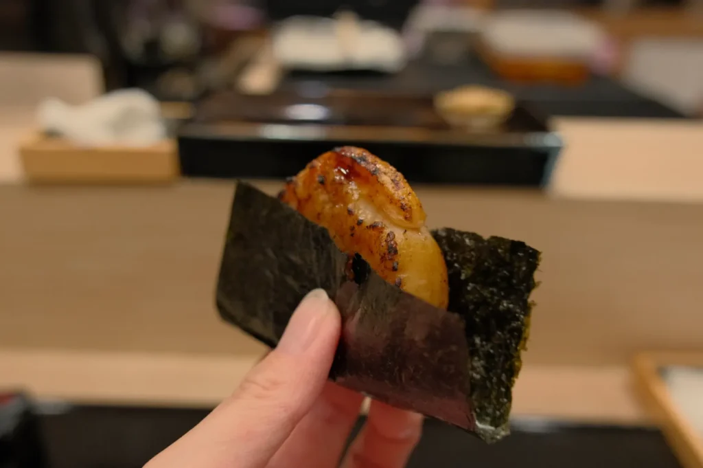 A hand holding a piece of sushi wrapped in nori with a piece of grilled fish on top.
