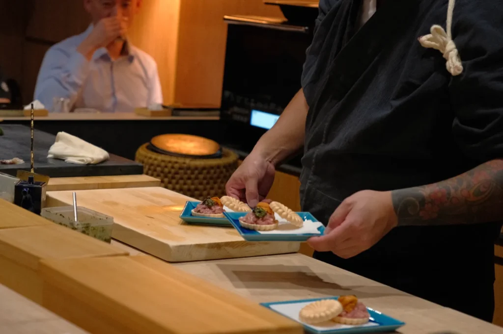 Chef Masa serving two small plates of food at a sushi bar, with a person in the background watching.
