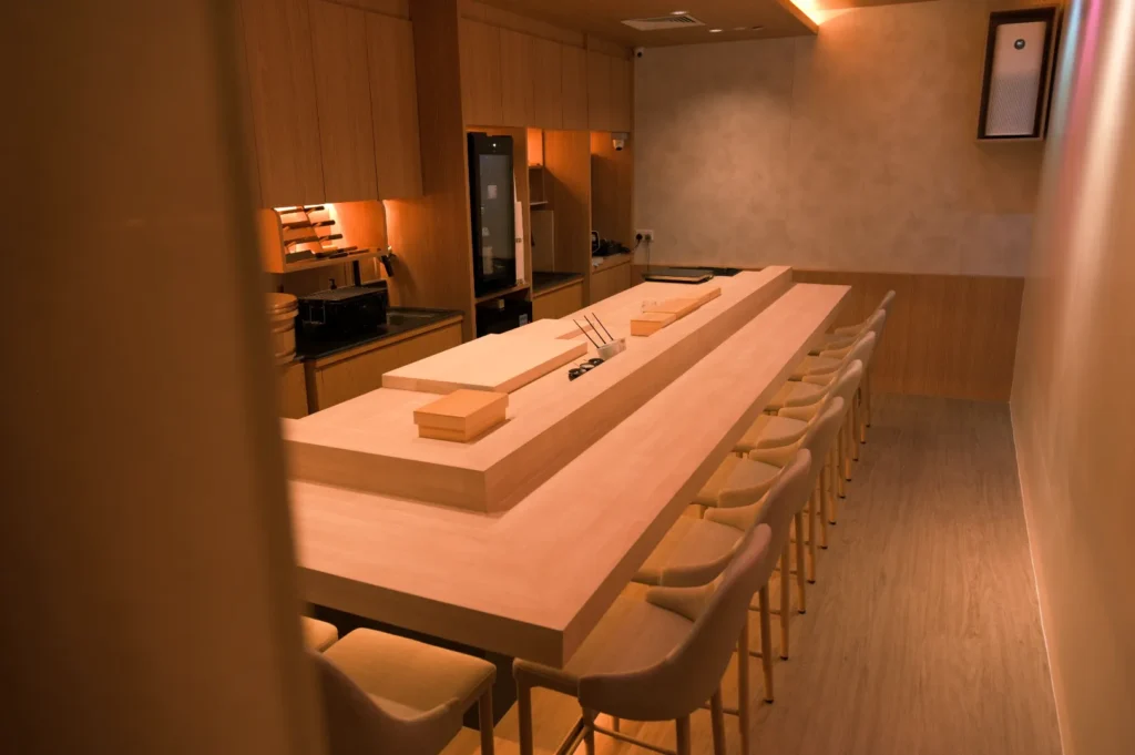 A minimalist kitchen interior with a large wooden table surrounded by chairs, featuring a sushi preparation area and modern appliances.