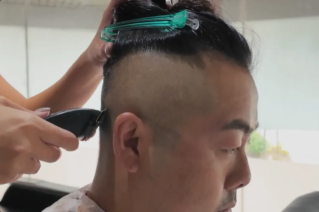 Chef Masa getting a haircut with the sides of his head shaved, while someone uses clippers on the back. He has a hair clip holding up the top section of his hair.