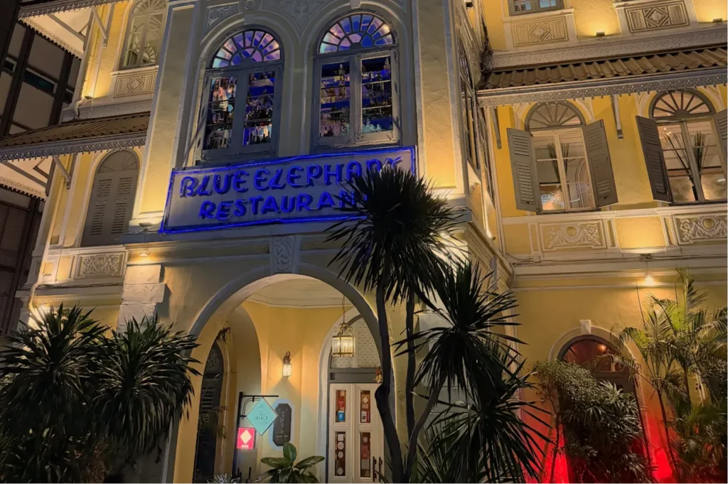 A brightly lit restaurant facade at night with the name "Blue Elephant Restaurant" prominently displayed, surrounded by palm trees and decorative architecture.