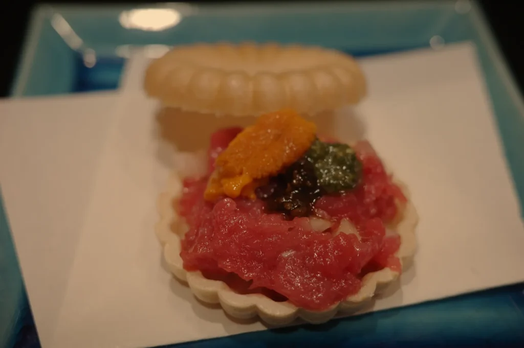 A small seafood dish served on a shell-like cracker, topped with raw fish, sea urchin, and a dollop of green sauce, presented on a blue plate.