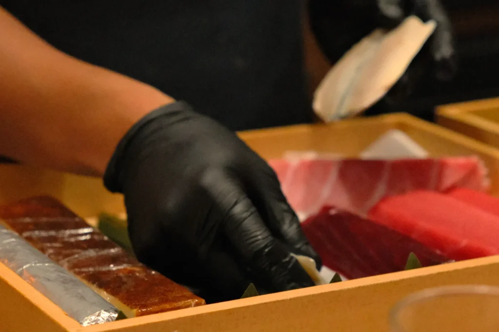 Chef Masa wearing black gloves is picking up a piece of sushi from a wooden tray containing various types of fish and sushi ingredients.