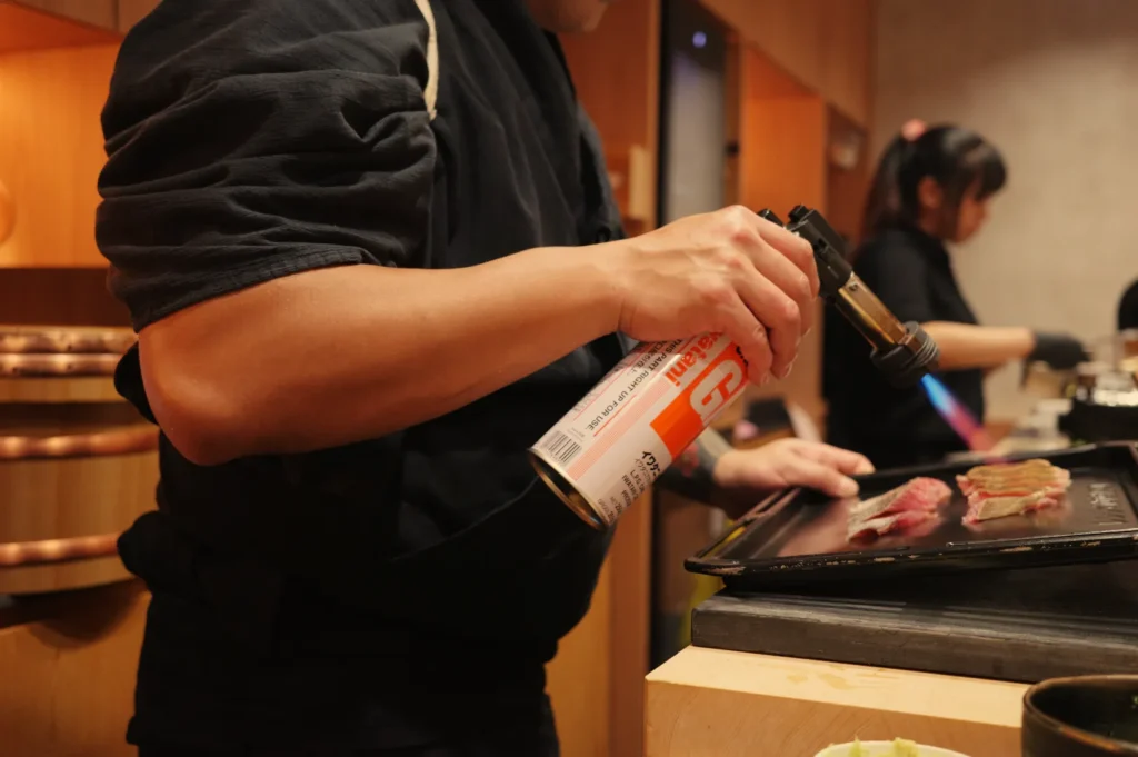 Chef Masa using a torch to sear meat at a sushi restaurant, with a kitchen helper in the background.