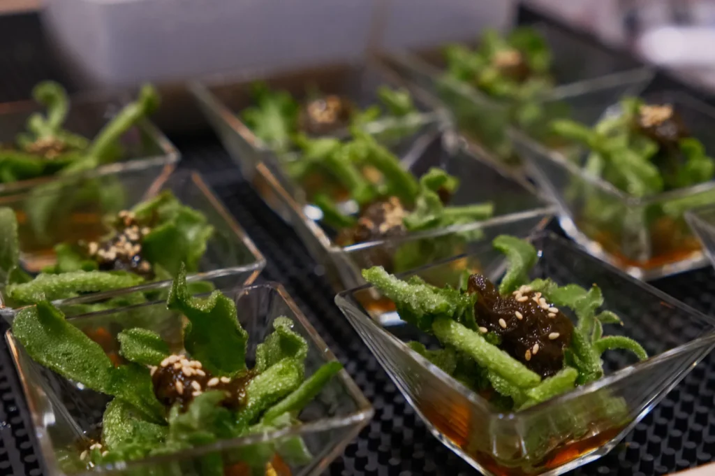 Small servings of green leafy vegetables topped with sesame seeds and a dark sauce, arranged in clear glass dishes.