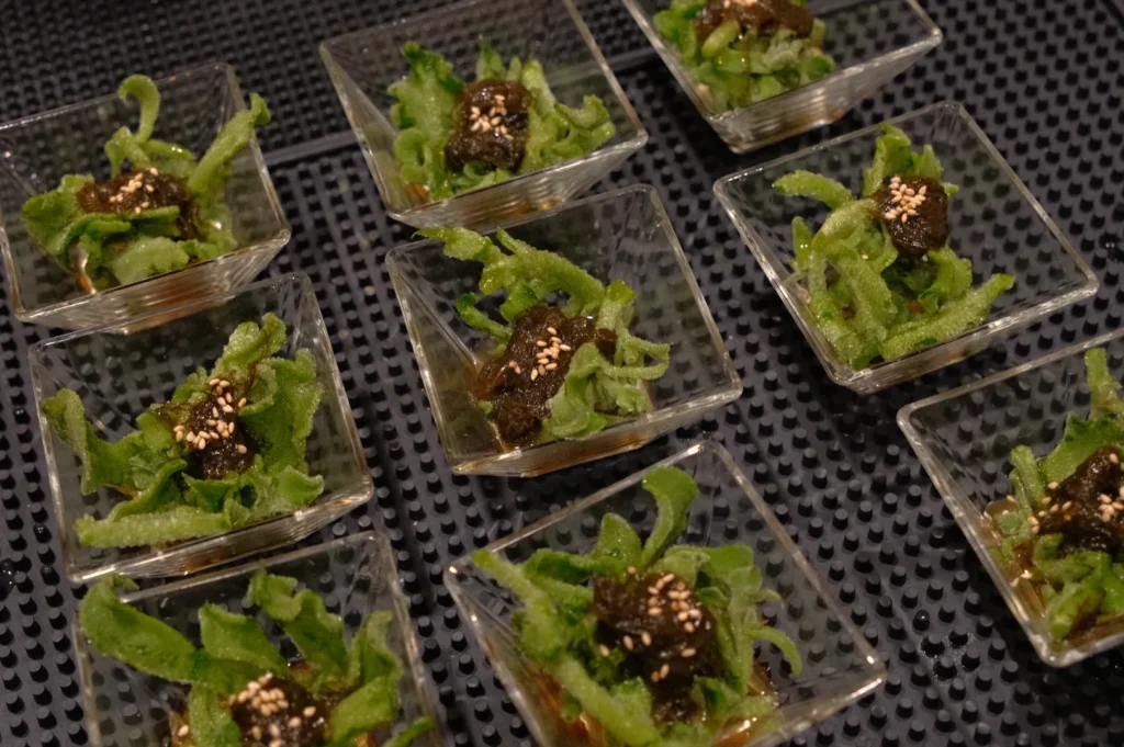 A close-up of small glass dishes containing green leafy vegetables topped with dark sauce and sesame seeds.