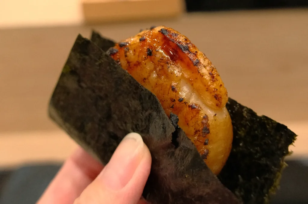 A piece of sushi, specifically nigiri topped with grilled seafood, held in a hand wrapped in seaweed.