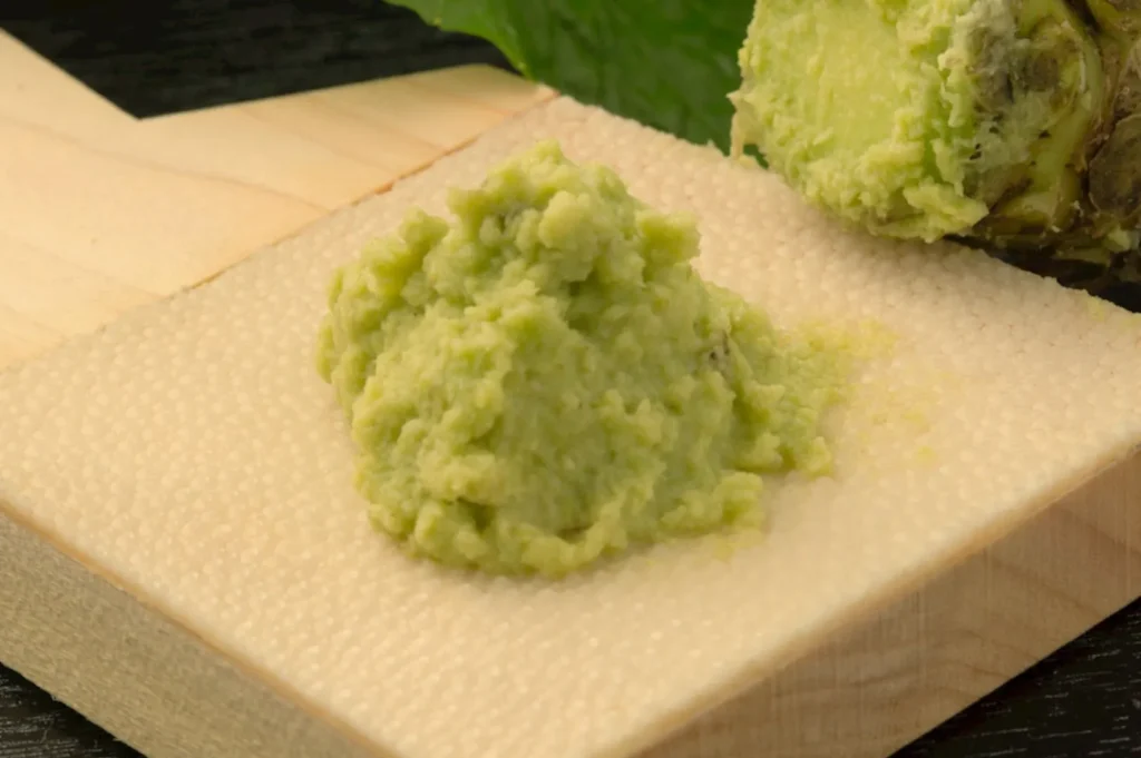 A mound of wasabi paste on a textured wooden surface, with a wasabi root in the background.