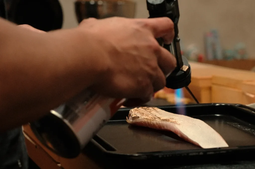 A close-up of a Chef Masa’s hand using a torch to sear a piece of fish on a black tray.