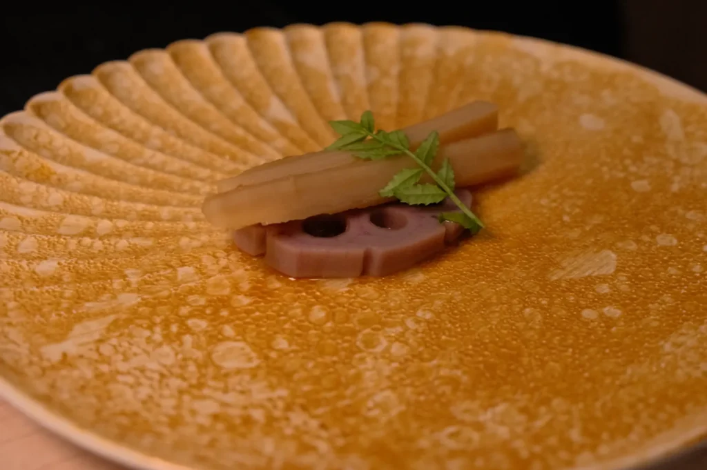 A plate with a decorative pattern containing a sauce, topped with pieces of root vegetable and garnished with a small green herb.