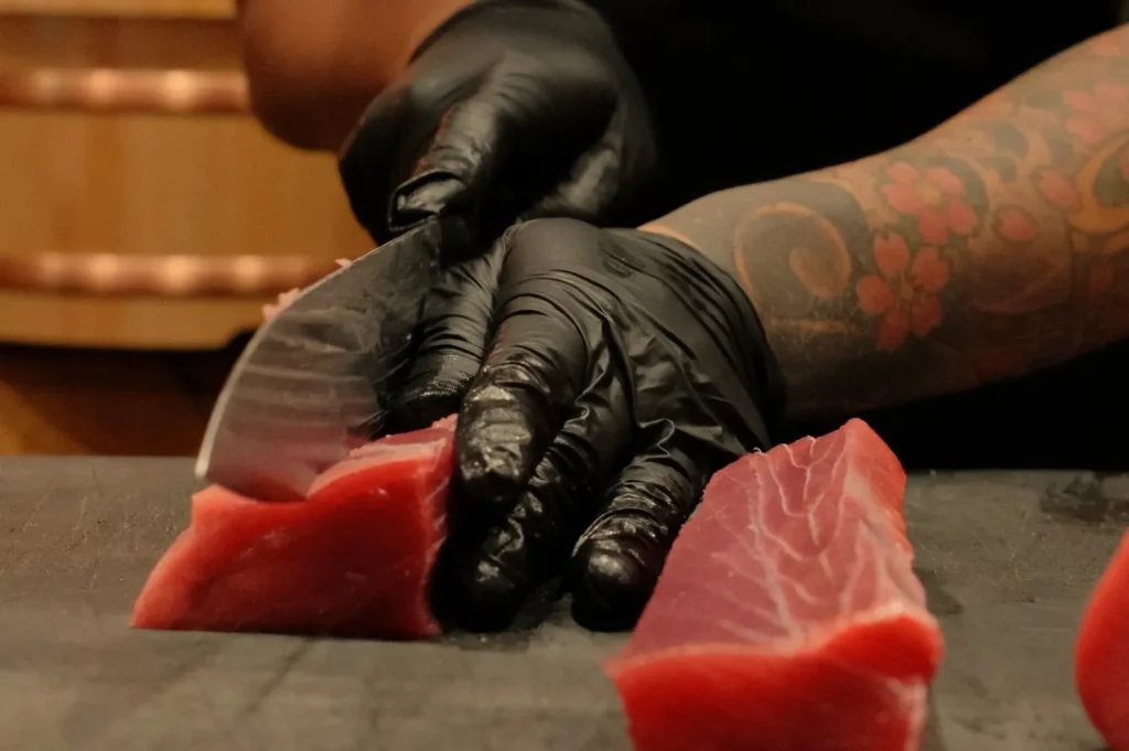 A person wearing black gloves is cutting pieces of raw fish, possibly tuna, on a cutting board. Their arm is adorned with colorful tattoos.