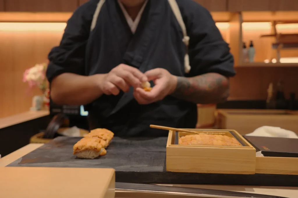 Chef Masa in traditional attire prepares sushi in a modern kitchen setting, with sushi ingredients arranged on a counter.