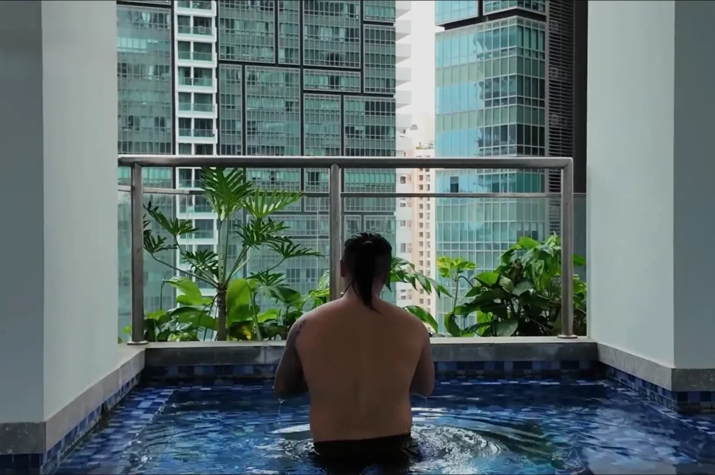 Chef Masa sitting in a small pool on a balcony, overlooking a cityscape with tall buildings and greenery.