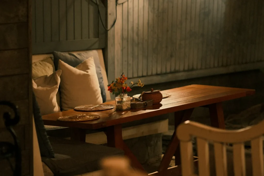 A cozy outdoor dining setting with a wooden table, decorated with flowers and soft cushions in a dimly lit environment.