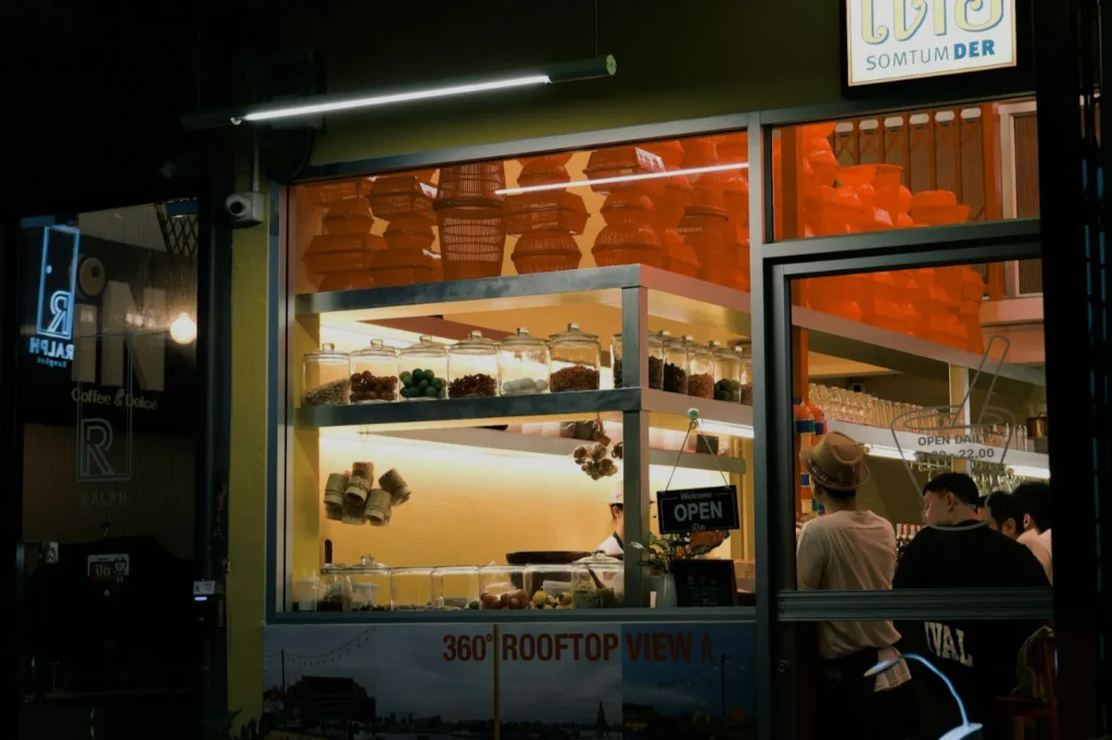 A view of a food stall or restaurant with an illuminated sign and visible shelves displaying food items. The interior is busy with customers, and the exterior signage indicates a rooftop view.