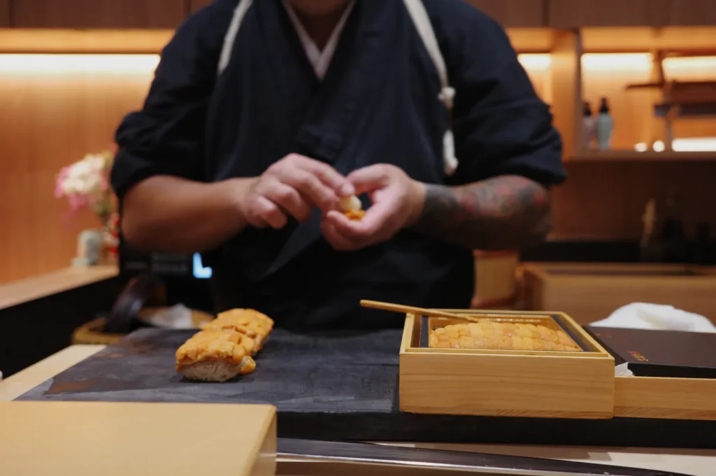 Chef Masa preparing sushi, focusing on a piece of sushi in front of him, with sushi on a wooden platter and a bamboo mat.