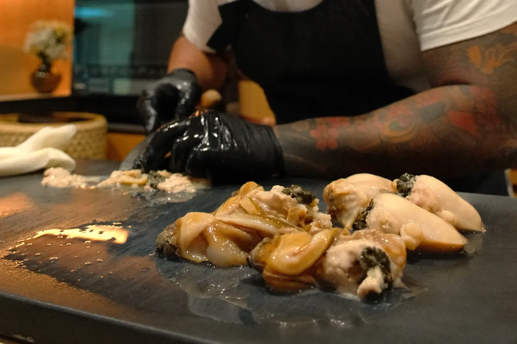 Chef Masa wearing black gloves is preparing seafood on a dark slate plate in a kitchen setting.