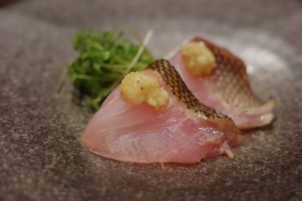 Two pieces of fish sashimi garnished with a dollop of sauce, accompanied by a sprig of microgreens, on a gray plate.