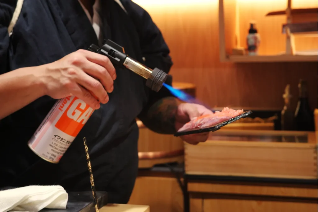 Chef Masa in a traditional attire using a torch to sear sushi or sashimi in a kitchen setting.