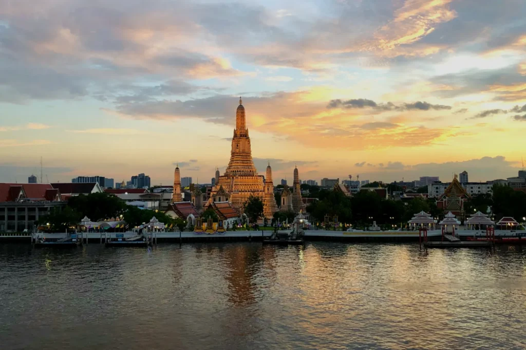 A view of Wat Arun temple during sunset, with the Chao Phraya River in the foreground reflecting the colorful sky.