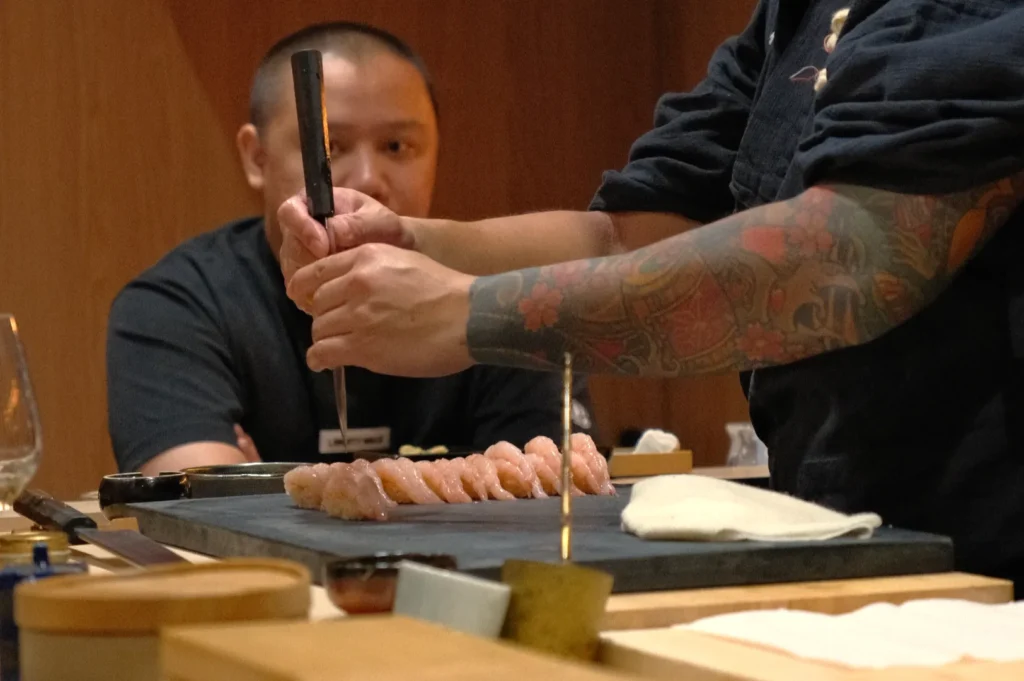 Chef Masa prepares sushi at a counter, with a focus on their hands and a selection of sushi in front. Another person is seated in the background, watching.
