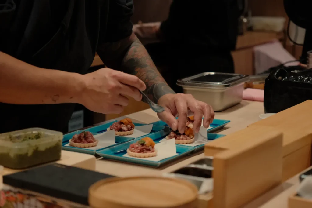 A chef prepares small dishes with toppings, using a spoon, in a restaurant kitchen.
