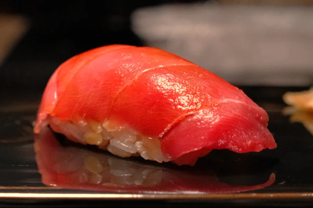 A piece of sushi featuring a slice of red tuna on top of rice, served on a dark plate.