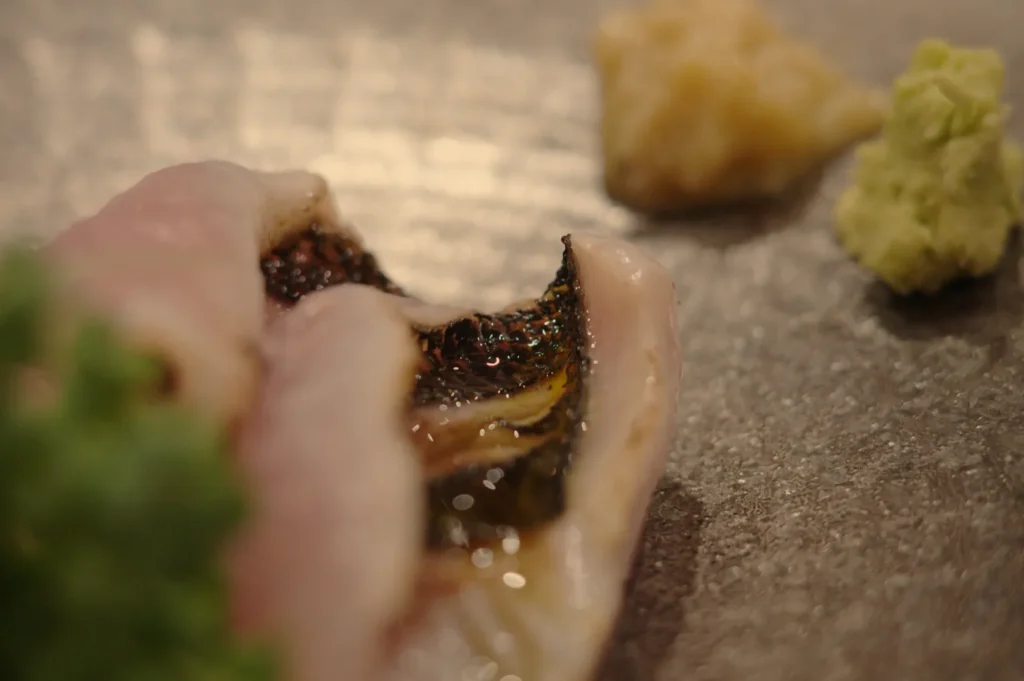 A close-up of a piece of sushi, featuring fish and garnishes like wasabi and ginger on a textured plate.