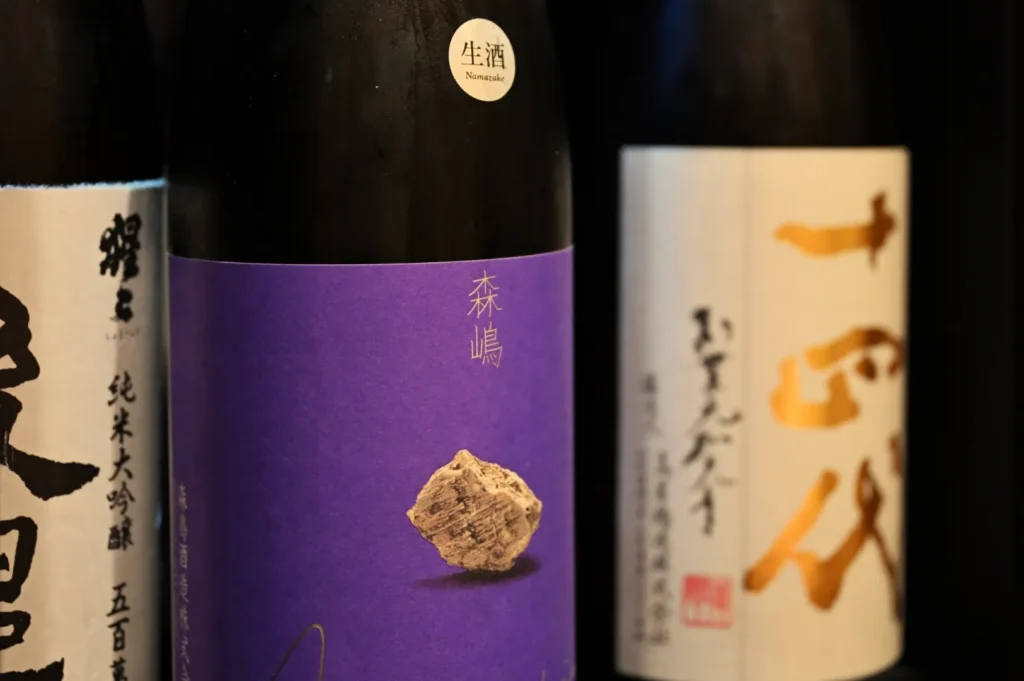 Three bottles of sake, with one featuring a purple label and an image of a stone, while the others have distinct labels in black and white.