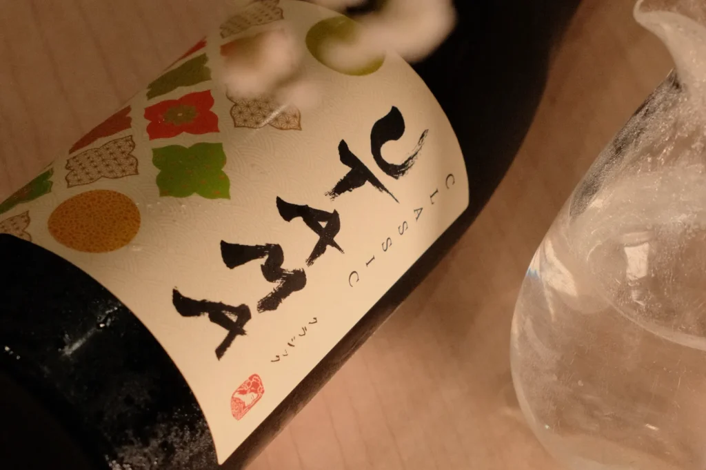 A close-up of a wine bottle with a unique label featuring various colorful designs and stylized text. A glass jug with water is also visible in the image.