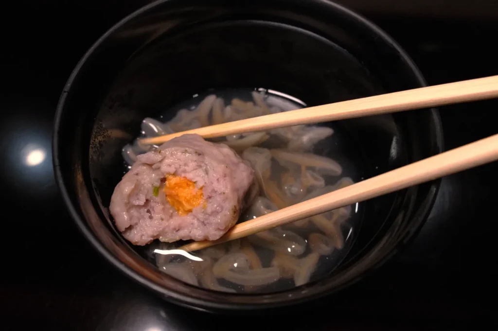 A bowl containing a translucent broth with a small dumpling topped with orange filling, being picked up with chopsticks.