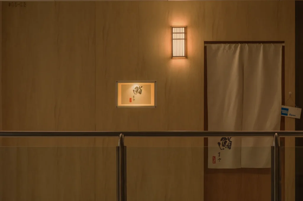 A softly lit hallway with wooden walls, featuring a wall-mounted light, a small illuminated square window, and a white curtain draped over a doorway.