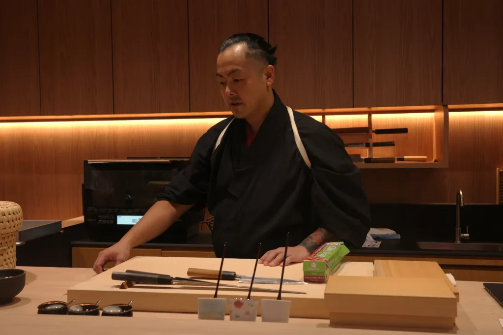 A chef in traditional attire prepares to work at a sushi counter, with knives and utensils laid out on the wooden surface.