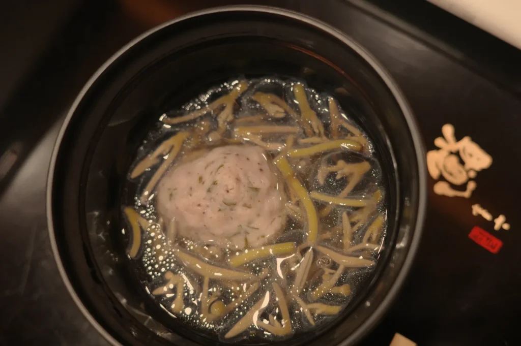 A round dumpling surrounded by thinly sliced vegetables in a dark broth, viewed from above.