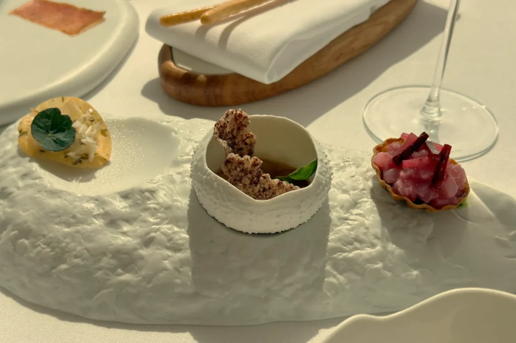 A gourmet dish presented on an artistic white plate, featuring small portions of food, including a pastry shell and garnishes, with a glass of wine in the background.