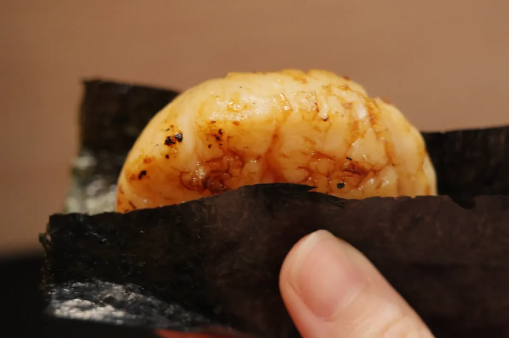 A piece of sushi with a grilled seafood topping, held by a person's hand and wrapped in seaweed.