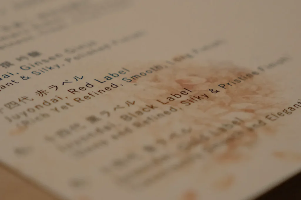 A close-up of a menu featuring wine options with descriptions, including "Juyondai: Red Label" and "Tsukasa Black Label." The background has a blurred, textured appearance.