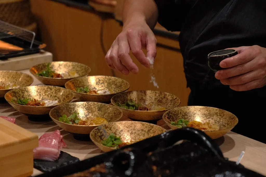 A chef preparing dishes in decorative bowls, sprinkling seasoning over them, with fresh fish in the foreground.