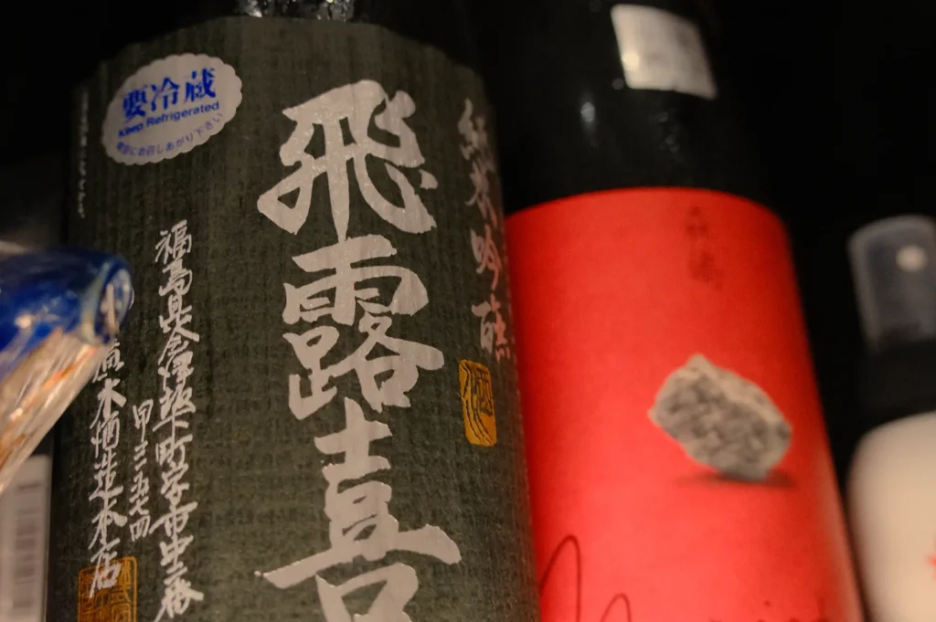 Two bottles of Japanese sake displayed on a shelf. One bottle has a dark label with white calligraphy, while the other has a red label with a simple design.