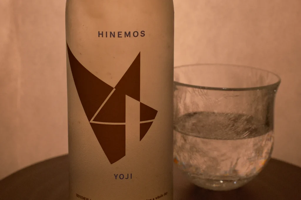 A bottle of Hinemos Yoji sake next to a glass of water on a wooden surface.