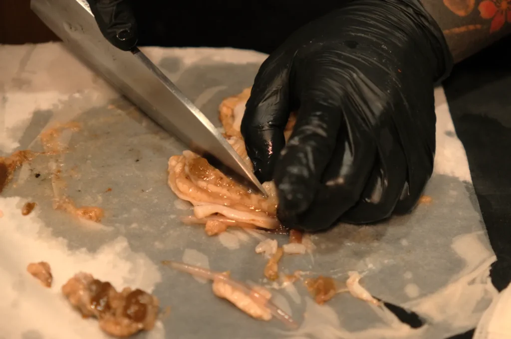 Chef Masa wearing black gloves is cutting raw meat on a piece of parchment paper with a knife.
