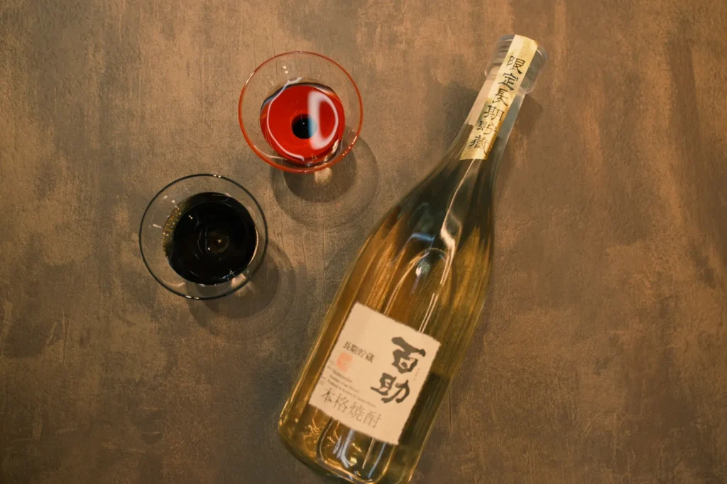 A bottle of sake with two glasses, one filled with red liquid and the other with dark liquid, placed on a textured surface.