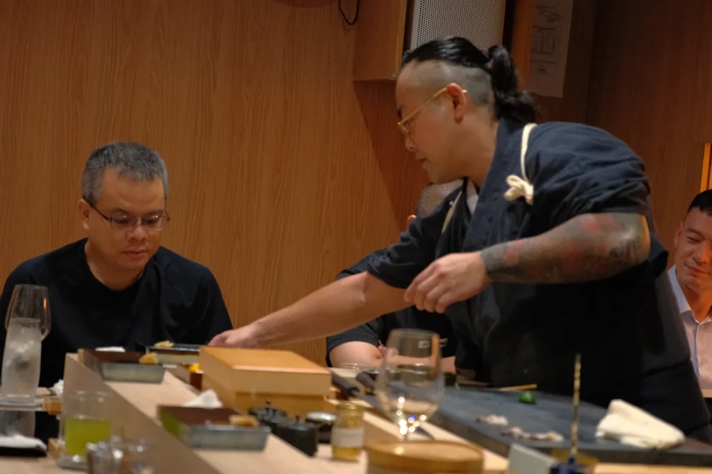 Chef Masa serving a dish to a diner at a sushi restaurant, with other diners in the background.