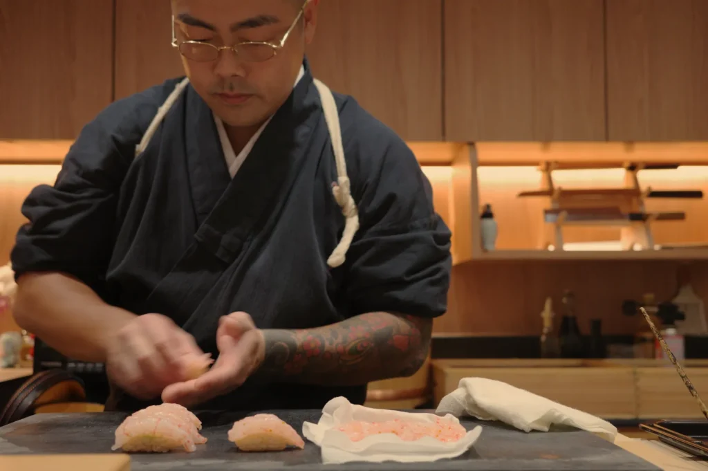 Chef Masa prepares sushi in a traditional setting, wearing a black outfit and glasses, with various sushi pieces displayed.